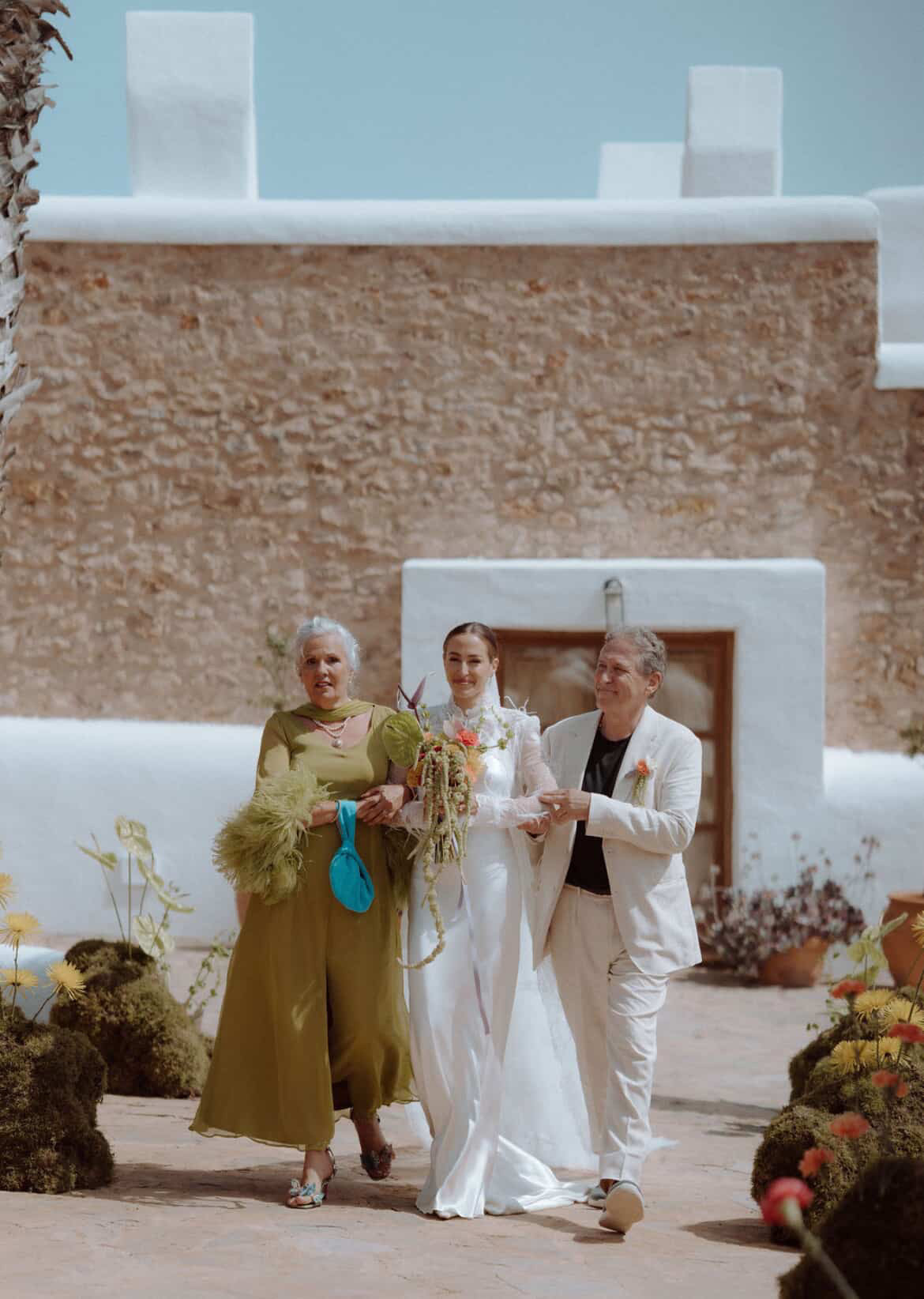 Three people standing together in front of a brick building with flowers and a clear sky.
