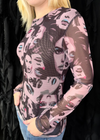 A woman wearing a sheer long-sleeve top with a pink, black, and gray graphic face print, paired with blue jeans, standing in front of a black draped background.
