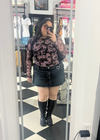 A person taking a mirror selfie in a clothing boutique, wearing a long-sleeve black and pink patterned top, a black mini skirt, and black knee-high boots.