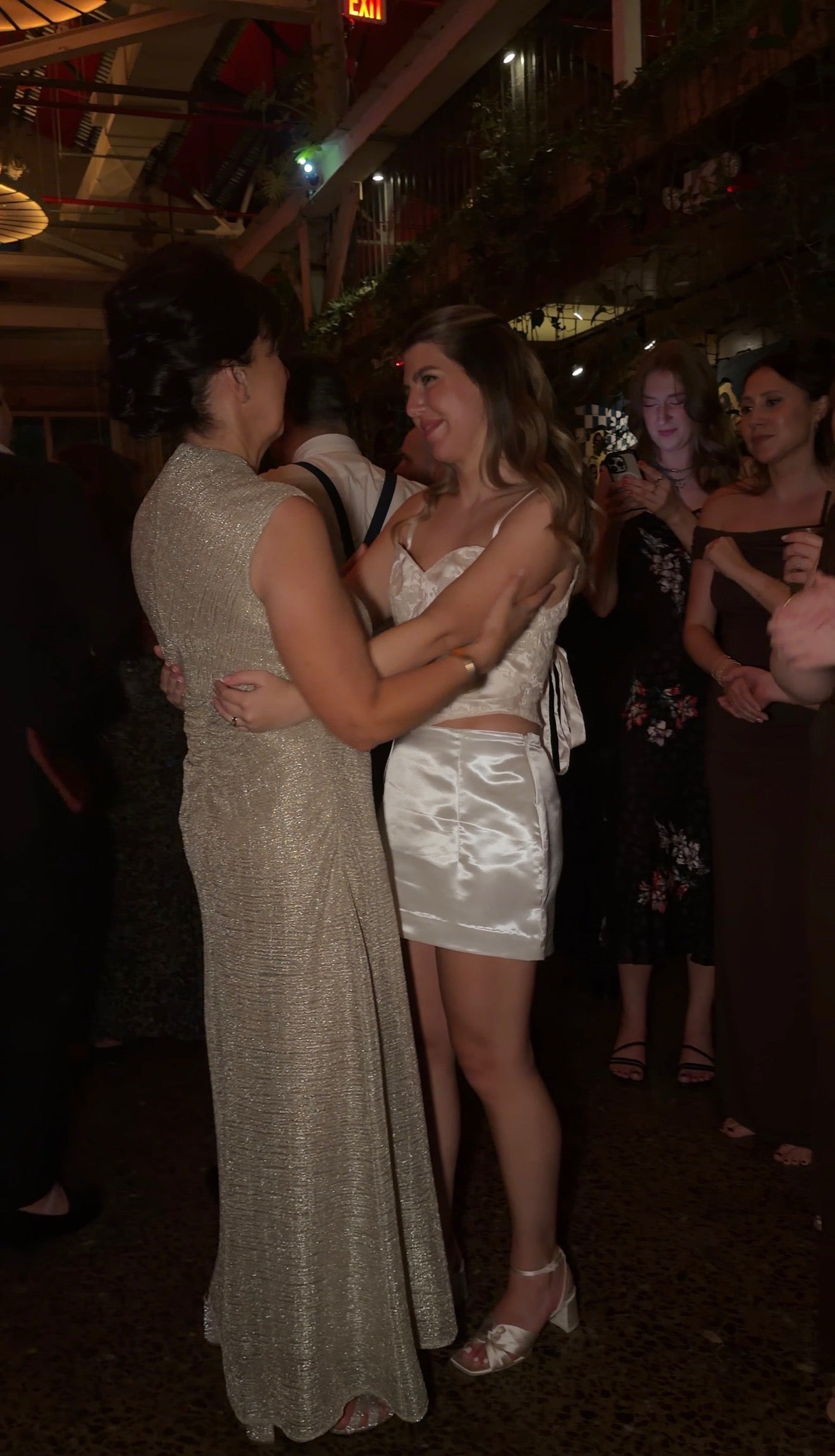 Two women embracing at a social event with other attendees in the background.